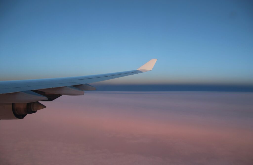 Image is of an airplane wing with blue skies above and pink clouds below, representing the safety habits needed before an international flight. 